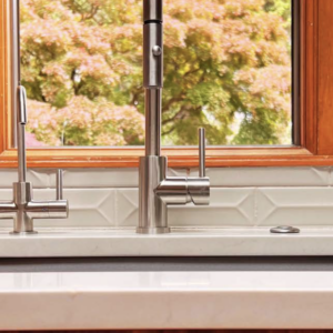 A modern kitchen sink with a stainless steel faucet set against a geometric-tile backsplash. Large windows provide a view of green trees. A roll of paper towels stands on the right side of the white counter.